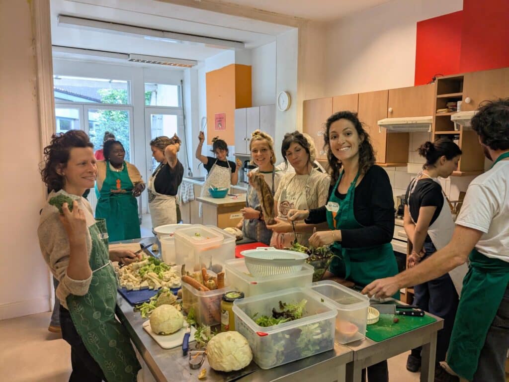 Photo montrant un groupe de personnes (majoritairement des femmes blanches dans leur trentaine) en cuisine, elles portent toutes un tablier. Il y a 4 femmes autour du plan de travail recouvert d'ingrédients qui sourient de face. Sur la droite, un homme est de côté et regarde l'espace cuisson. Au fond, deux femmes discutent et une autre lève un fouet au dessus de sa préparation.