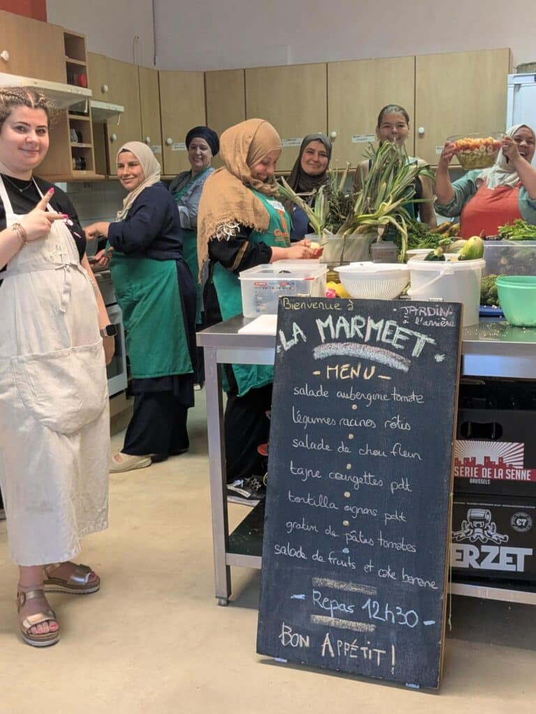 Image d'un groupe de 7 femmes de différents âges en train de cuisiner. 5 portent le voile et l'une d'elle porte une salade en l'air en riant. Au premier plan, la plus jeune fait le signe de la paix avec ses mains.